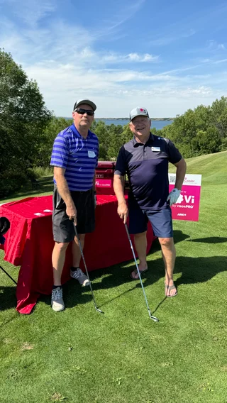 John (Left) standing next to Rob (Right) smiling directly in front of the camera while holding golf clubs standing on a course in front of a table decorated with Vi branding.