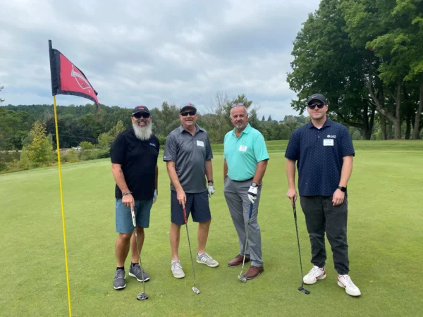 Vi Industry Consultant, John Schmisner, standing next to three other individuals next to a hole on the golf course.